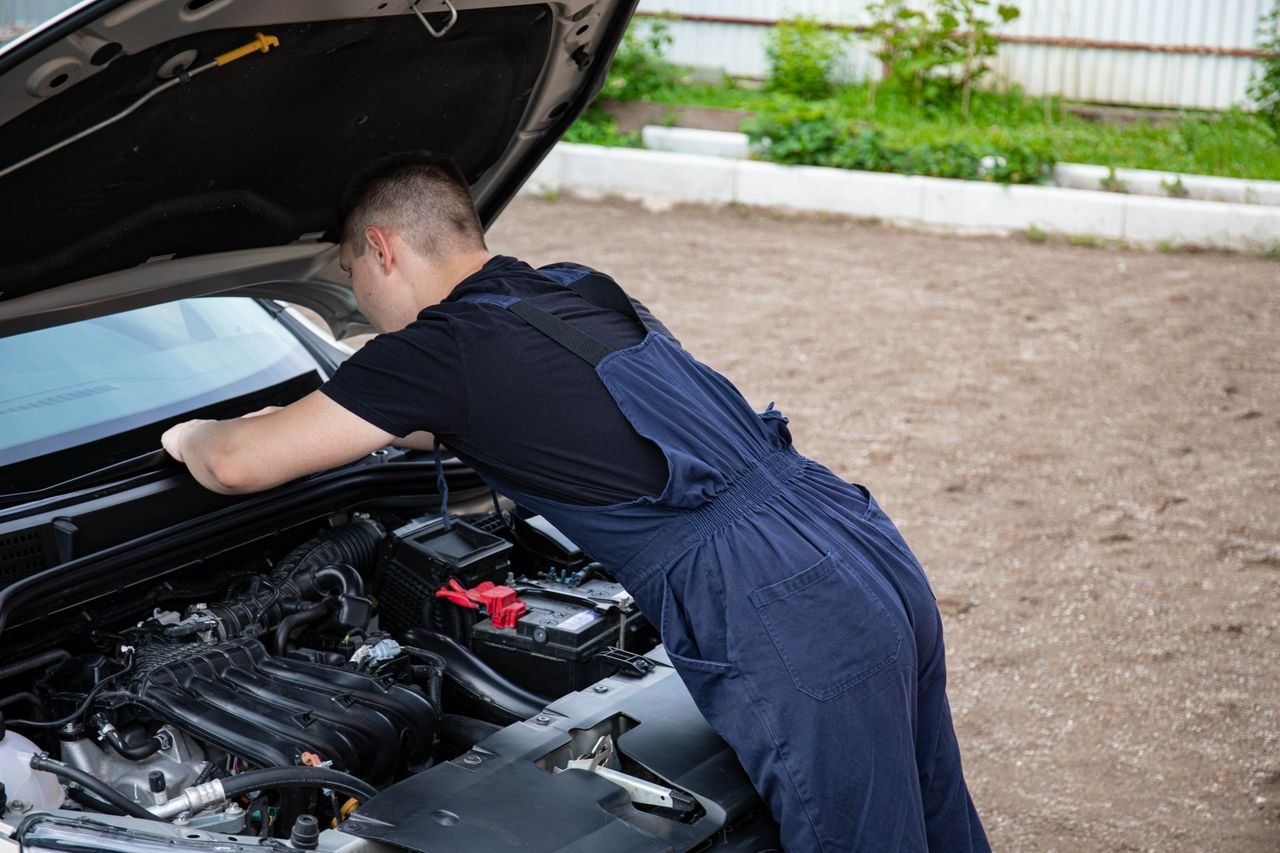 services-09 Mechanic in blue coveralls working on a car engine outdoors, focused on repair.